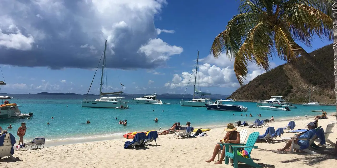 A white-sand beach with sunbathers and palm trees, catamarans and sailboats anchored in clear turquoise water beyond