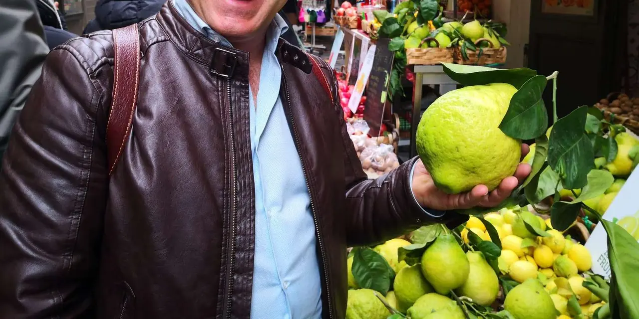 A guide in a brown leather jacket holding up a large lemon at a produce stall in a Catania market