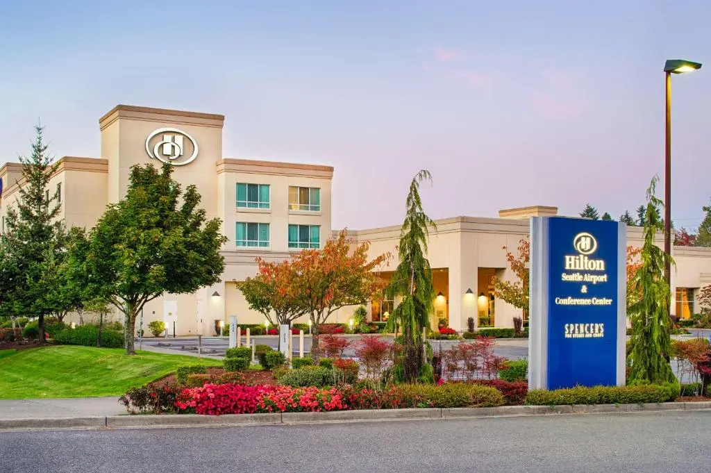 The cream-coloured Hilton Seattle Airport & Conference Center at dusk behind flower beds and its blue sign