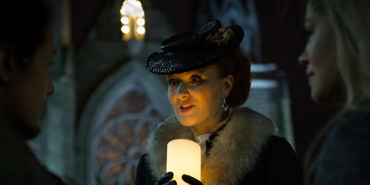A woman in a dark feathered hat and fur collar holding a lit candle as she talks to listeners at night