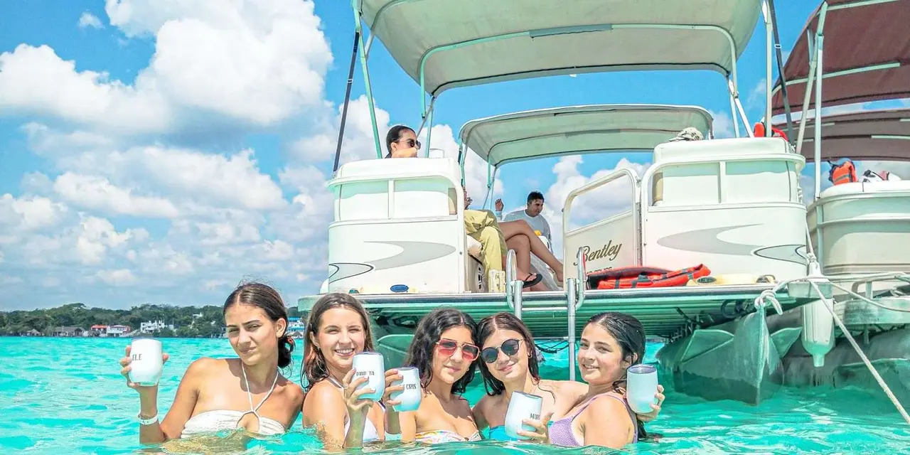 Five swimmers holding cups in turquoise lagoon water beside a canopied pontoon boat, with a green shoreline behind