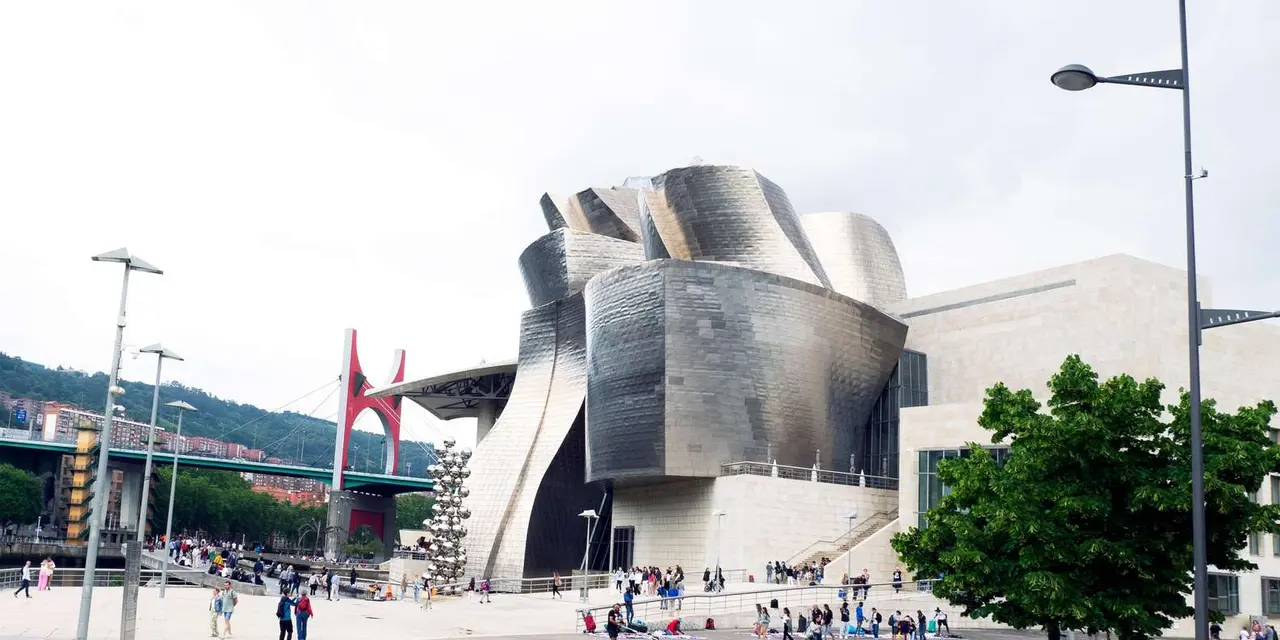 The Guggenheim's curved metal walls with the red arch of a bridge and visitors in the plaza under a pale sky