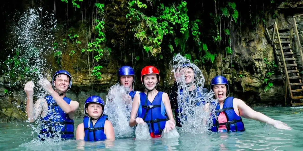 A group in helmets and life jackets splashing and cheering in a pool beneath jungle-covered rock