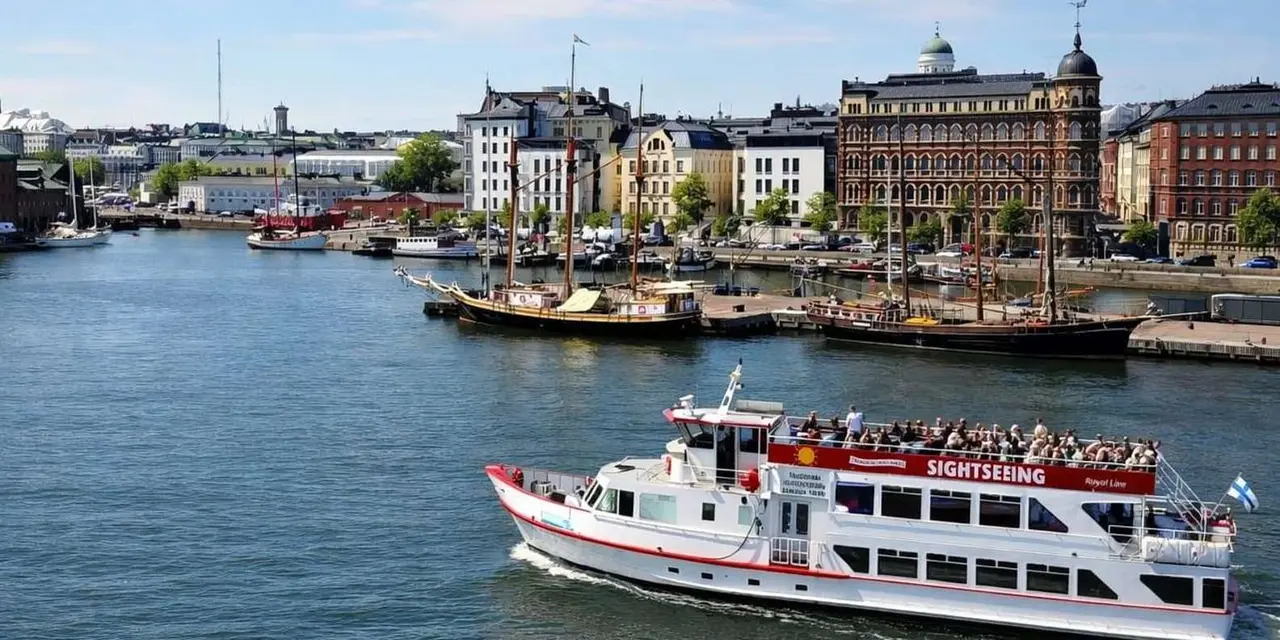 A white and red sightseeing boat crossing the harbour with passengers on deck and old sailing ships moored along the waterfront behind