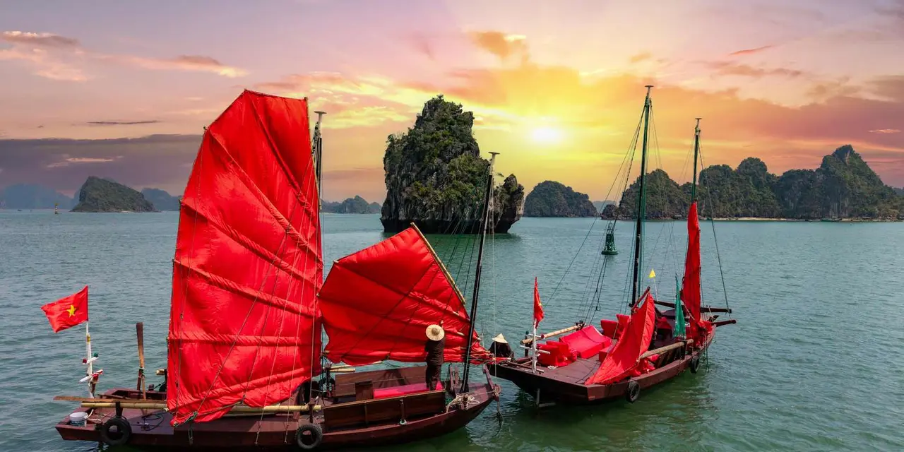 Two wooden boats with bright red sails against a limestone islet at sunset