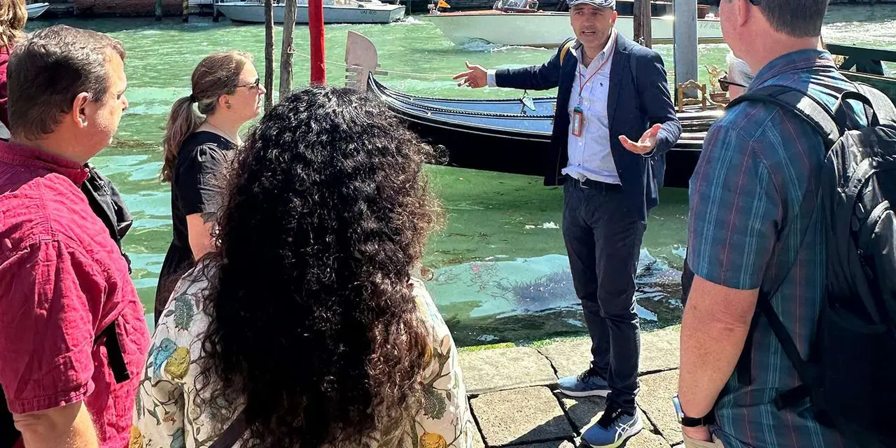 A guide talking to a small group on a waterfront by the Grand Canal with gondolas moored behind