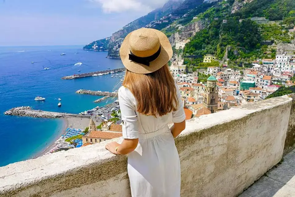A woman in a straw hat looking down over Amalfi's harbour, rooftops and the green coastline