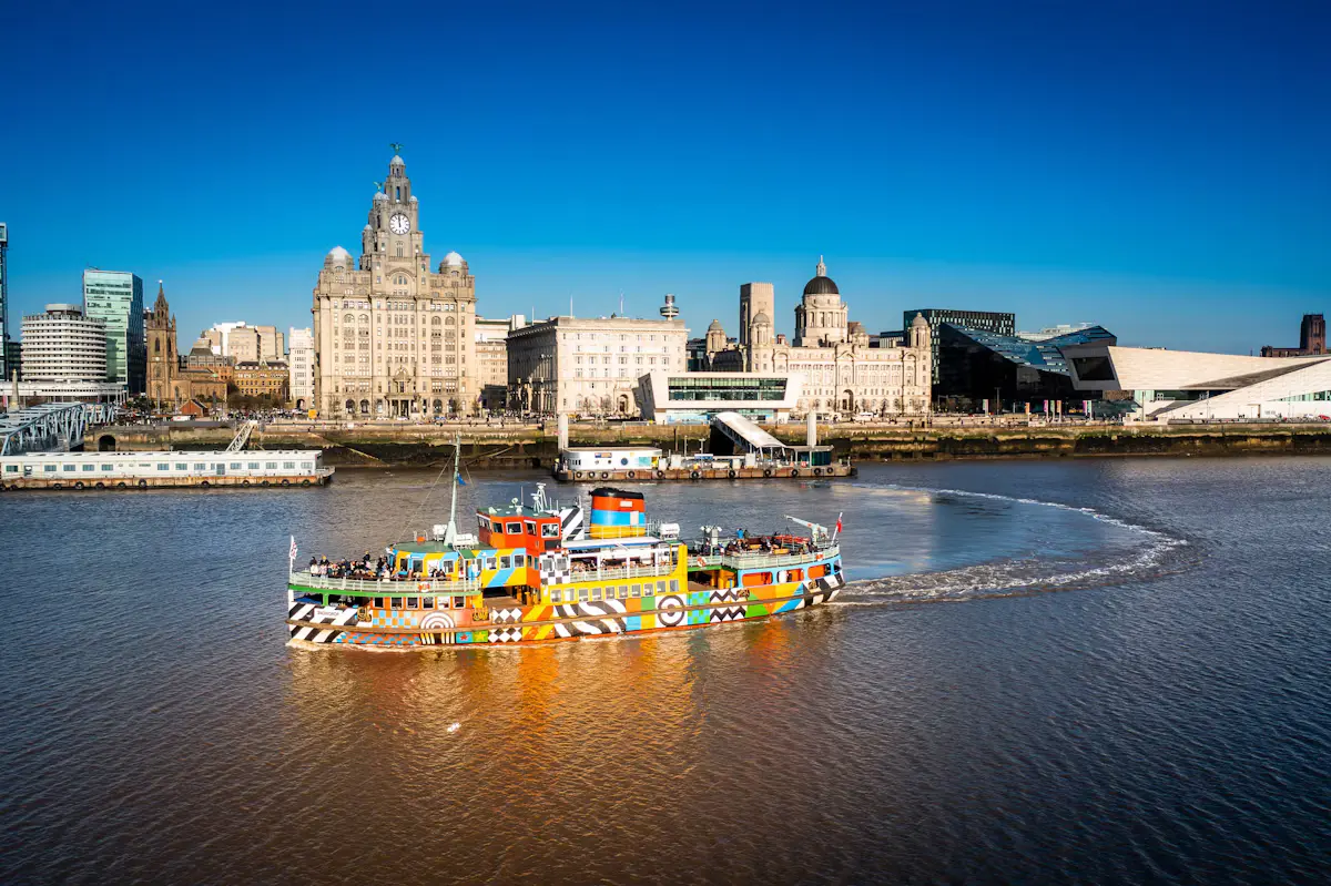 A brightly patterned Mersey ferry crossing the river with the Royal Liver Building and the Pier Head skyline behind it under a deep blue sky