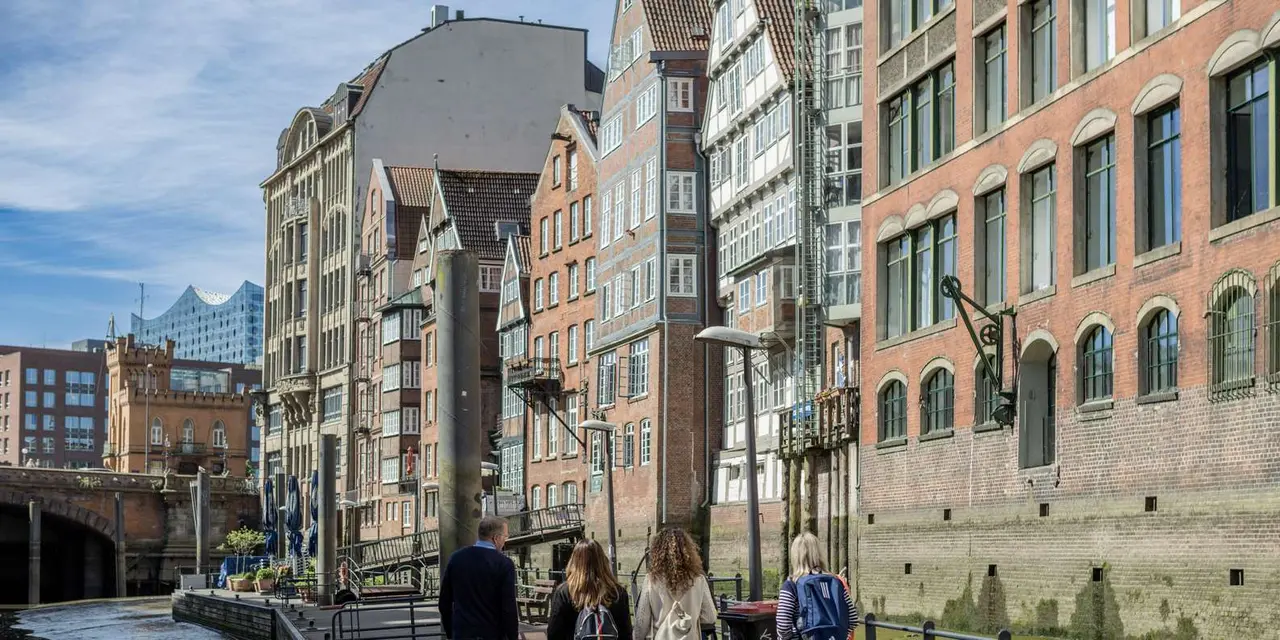 Four walkers heading along a canal-side path past narrow Hamburg brick houses, with the Elbphilharmonie's glass roof visible behind them