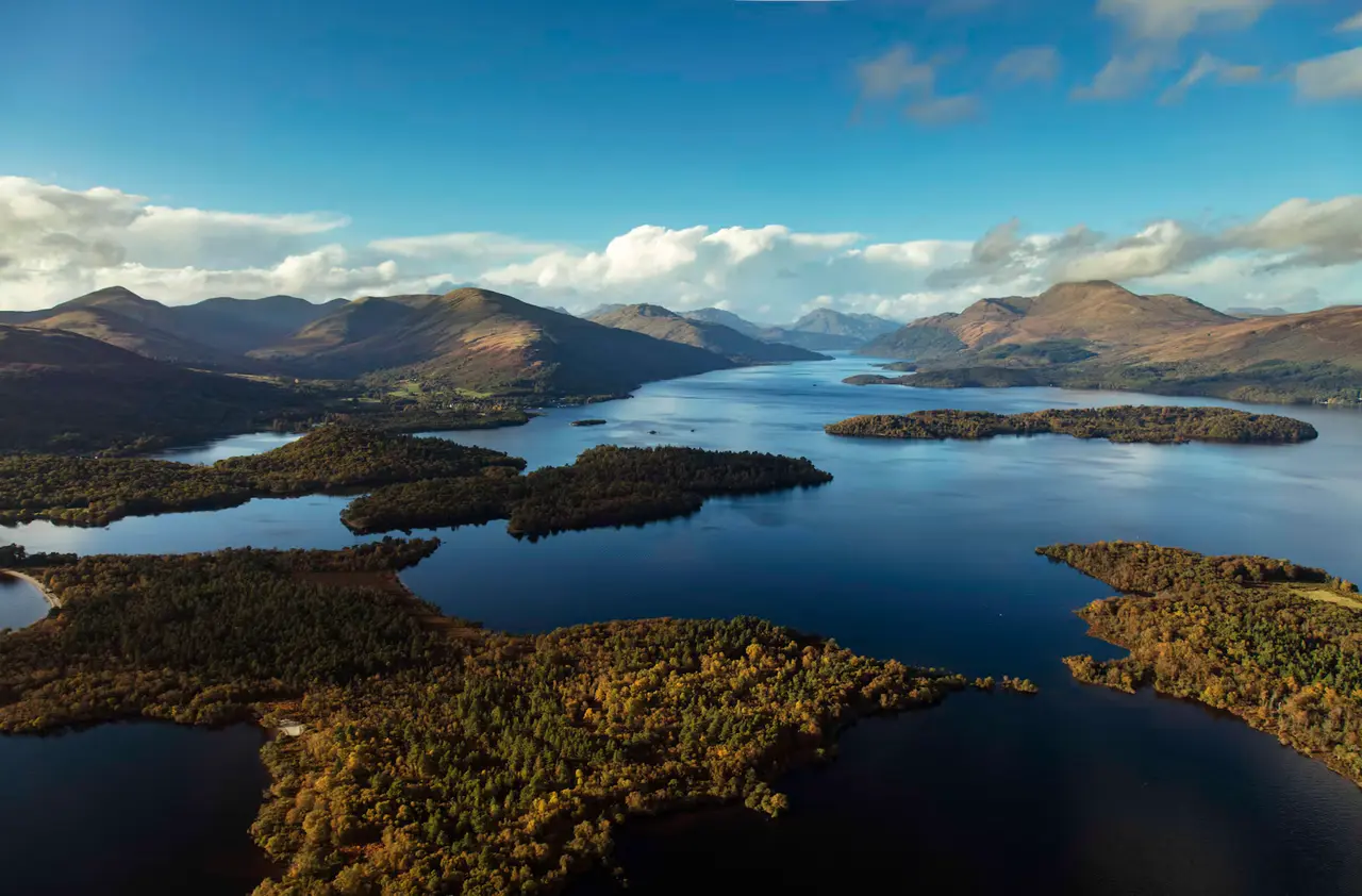 Aerial view of Loch Lomond's blue water and wooded islands, with rounded mountains and a band of cloud beyond — about 45 minutes from Greenock's cruise terminal