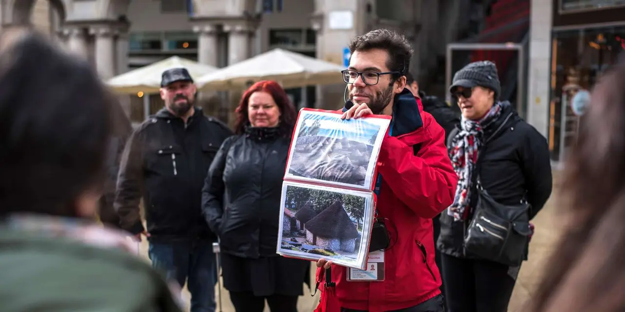 A guide holding up old printed pictures to a small group of visitors in coats on a city street