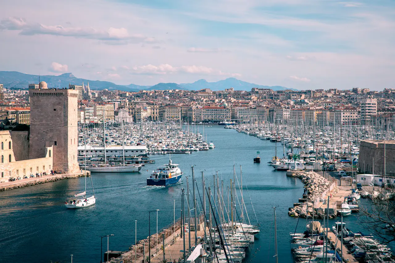Marseille's Vieux-Port seen from above its entrance, with the square stone tower of Fort Saint-Jean on the left, a ferry heading out between rows of yachts and the city climbing to the hills behind