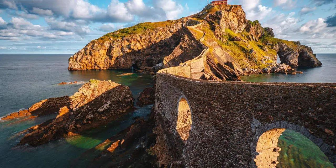 A stone bridge arching out to a rocky islet with a red-roofed chapel on top, sea and clouds behind