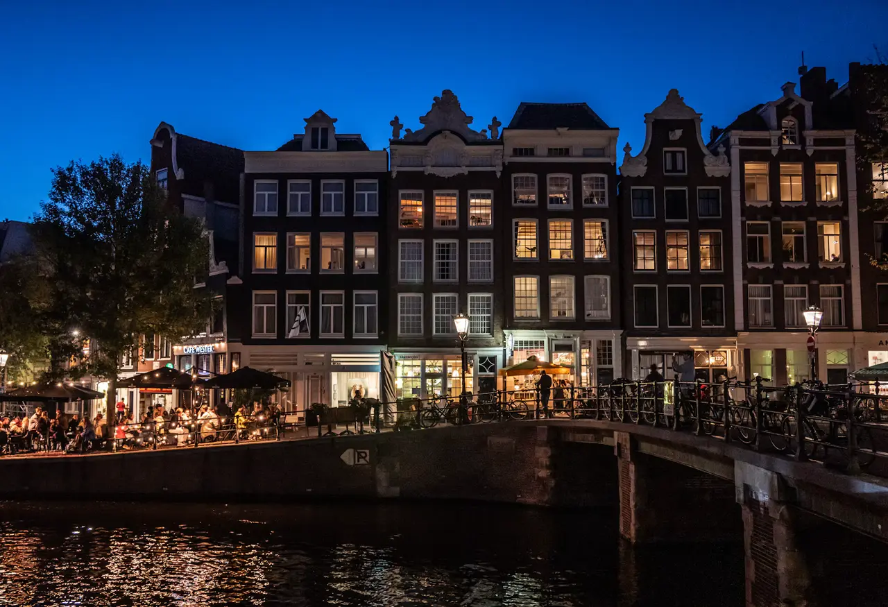 Tall gabled canal houses with lit windows beside a still canal at dusk, a pavement cafe with umbrellas on the left, bicycles on the bridge railing and a deep blue sky above