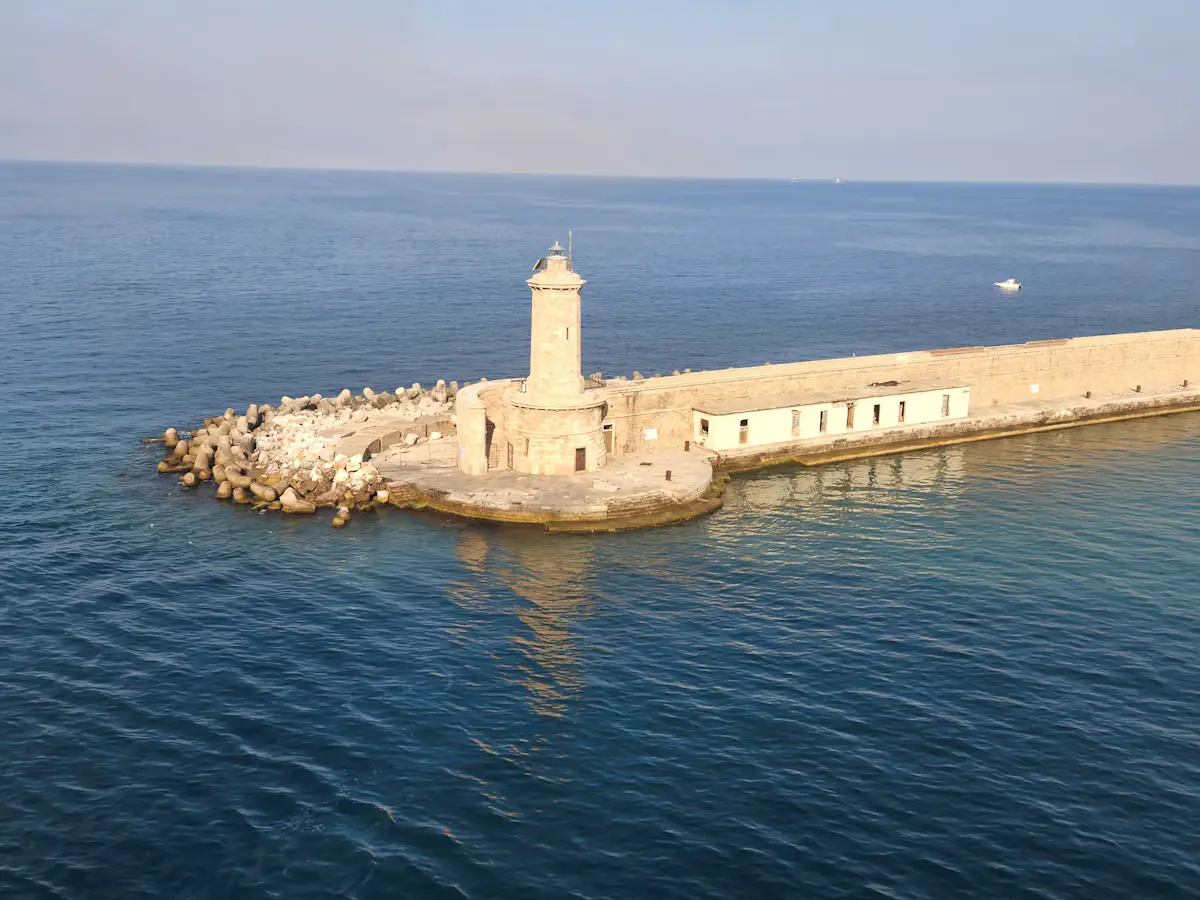 The stone lighthouse at the tip of Civitavecchia's harbour breakwater, seen from above on a calm, deep-blue sea in soft morning light