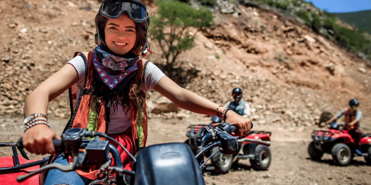 A smiling rider in a helmet and bandana sitting on an ATV on a dirt trail with other riders behind