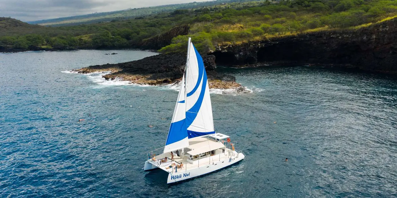 A white sailing catamaran under sail on deep-blue water off a dark lava coastline