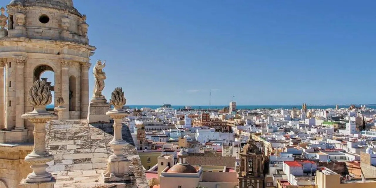 Stone finials and the corner of a bell tower on a rooftop, with the white buildings of Cádiz and a strip of sea behind