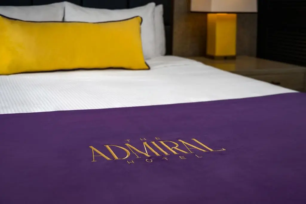 A bed at The Admiral with a purple runner embroidered with the hotel's name and a yellow pillow