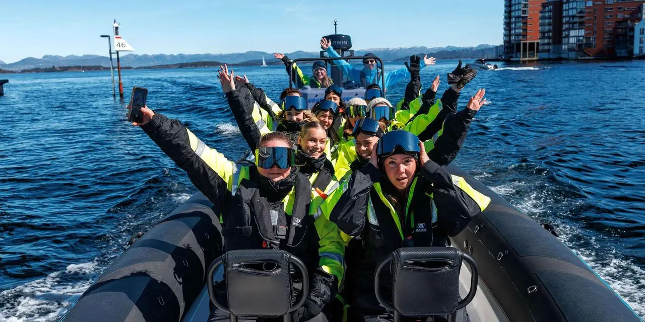 A boatload of passengers in flotation suits and goggles cheering with their arms in the air