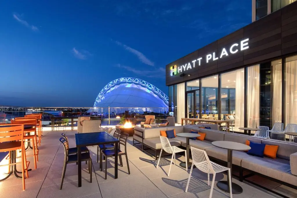 Hyatt Place Boston/Seaport's rooftop terrace at dusk, with a fire pit, orange cushions and the lit dome of the Seaport pavilion beyond