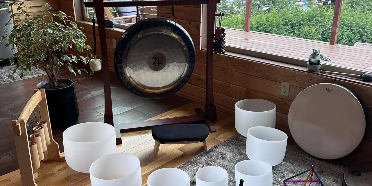 A large gong on a stand, white singing bowls and a frame drum on a wooden floor beside a window overlooking trees