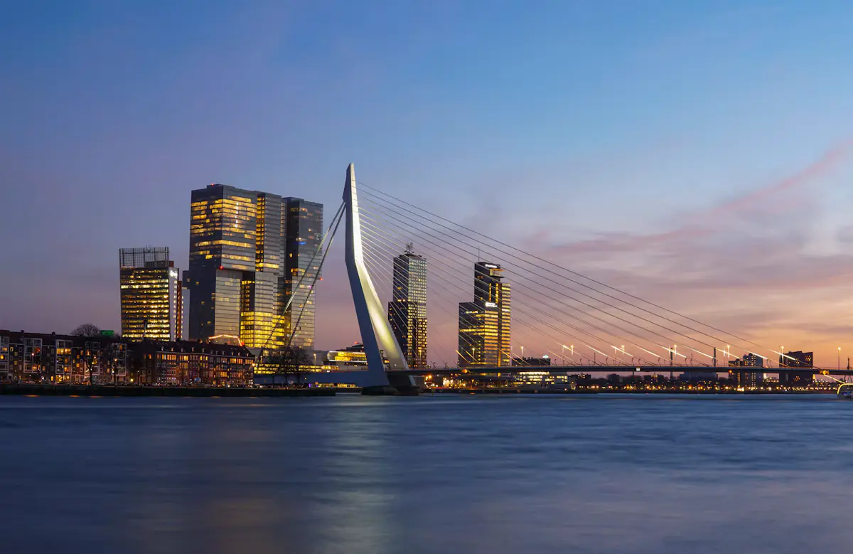 The Erasmus Bridge and the lit-up De Rotterdam towers on the Wilhelminapier across the Nieuwe Maas at dusk, under a blue-to-orange sky