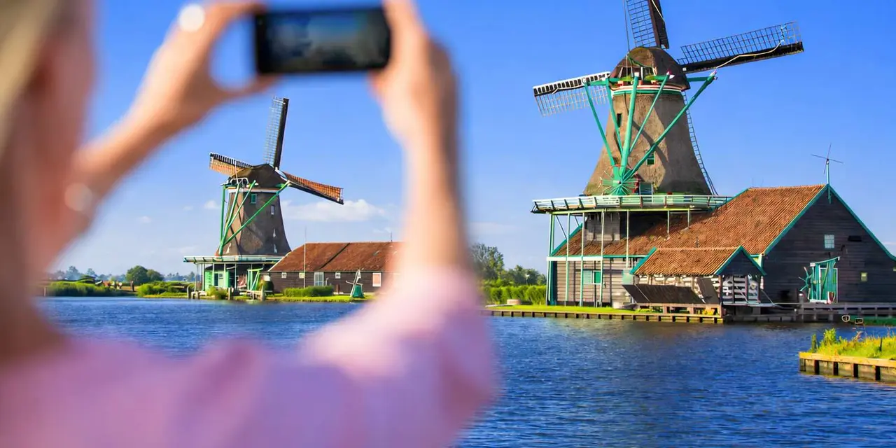 A hand holds up a phone to photograph two windmills with green and brown sails beside a blue canal under a clear sky