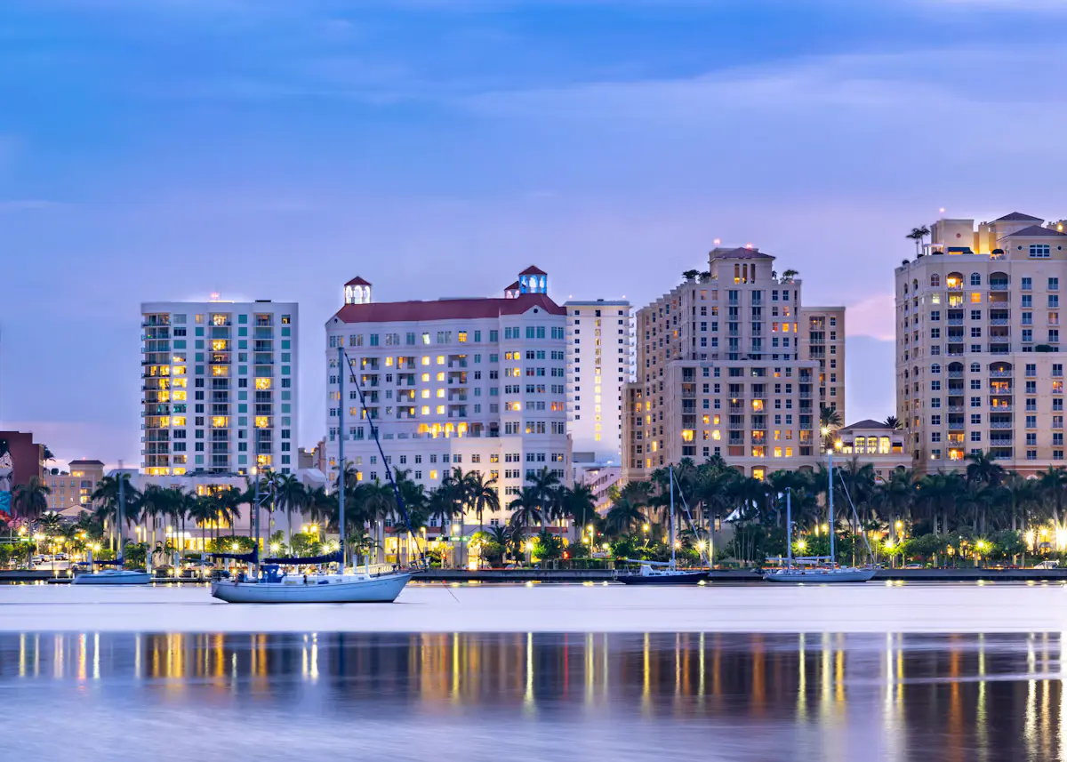 A sailboat anchored on the still water in front of lit-up waterfront apartment towers and palm trees in West Palm Beach at dusk