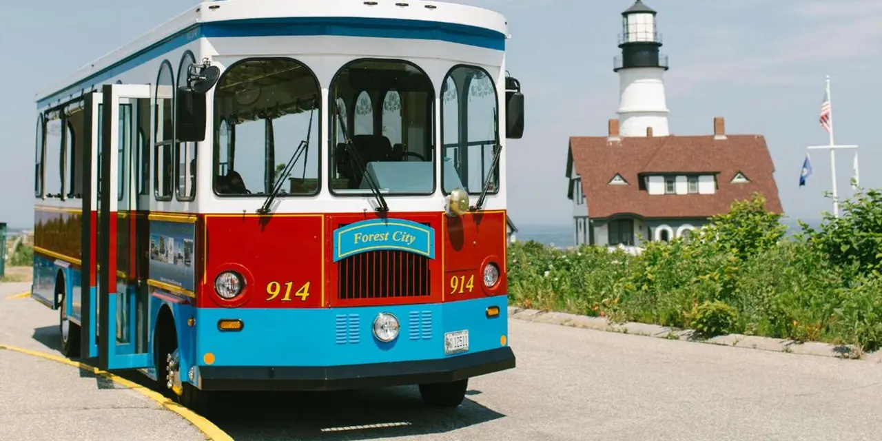 A red, white and blue trolley beside Portland Head Light and its red-roofed keeper's house