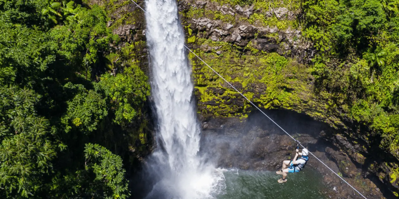 A rider in a harness ziplining across a green gorge beside a tall waterfall