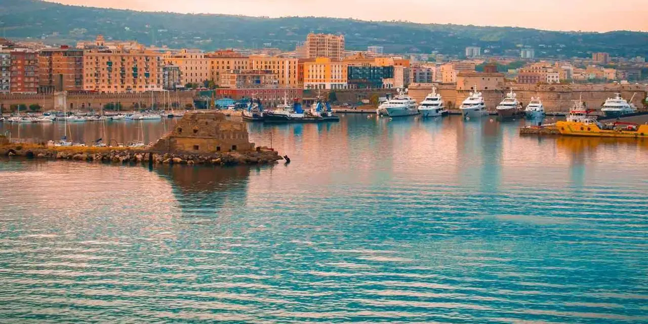 Civitavecchia's waterfront at dusk: pastel buildings along the harbor, boats in the marina and a small stone fort on the water
