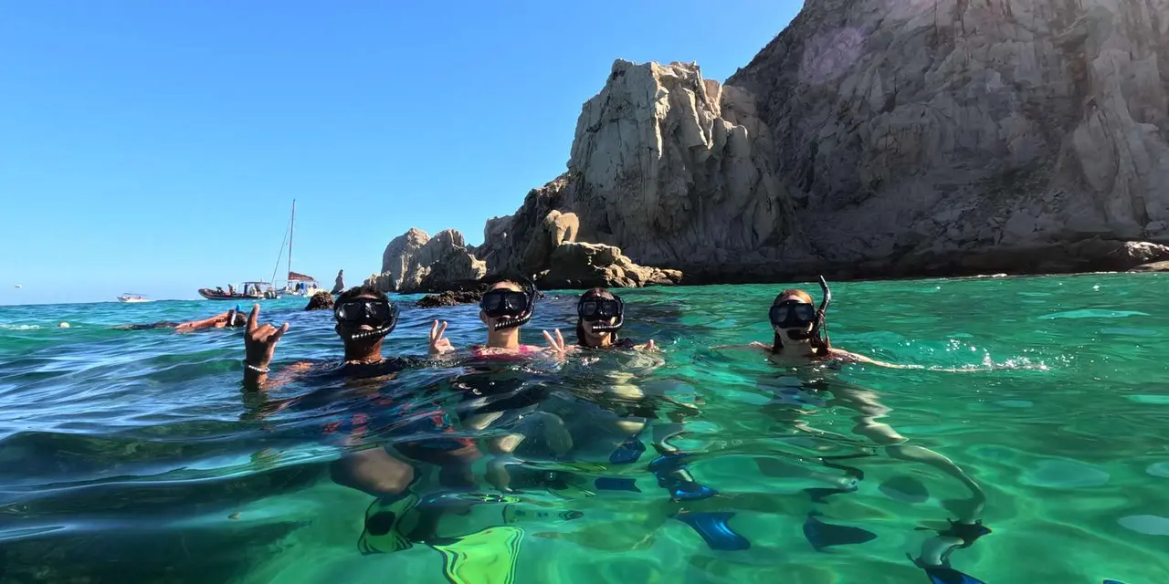 Snorkelers in masks floating in clear turquoise water below a tall rock headland