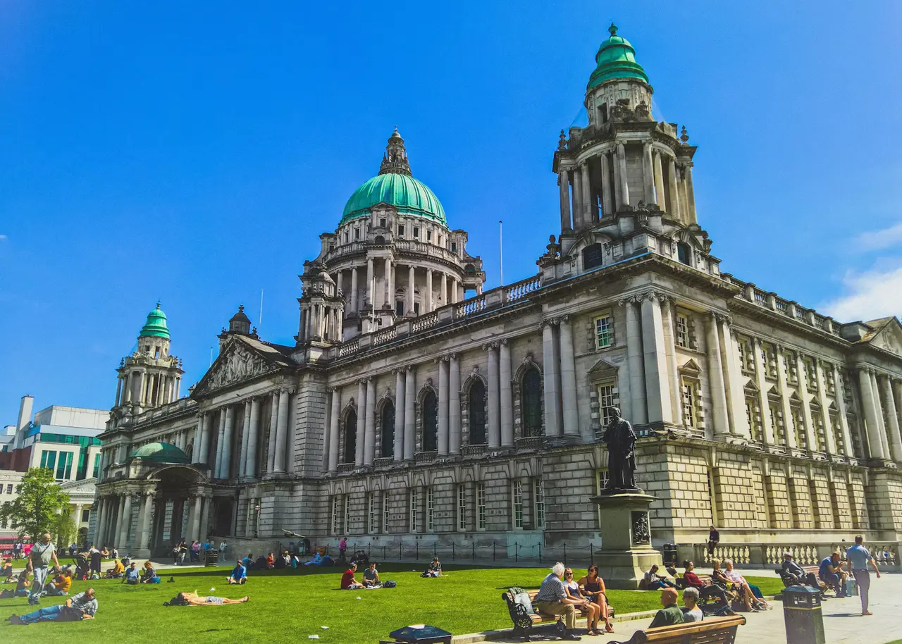 Belfast City Hall's green copper dome and corner towers in pale stone under a vivid blue sky, with people sitting on the lawn in the foreground