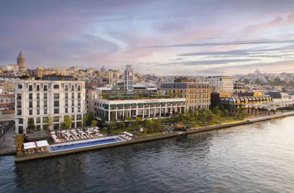 Aerial view of The Peninsula Istanbul's restored buildings on the Bosphorus at dusk, with a waterside pool and loungers in front and Galata Tower on the hill behind