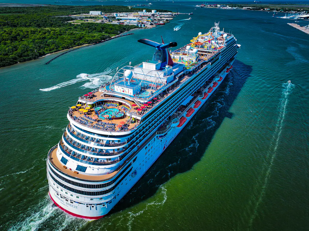 Aerial view of the Carnival Breeze sailing through green water off Galveston, its pool deck, red-and-yellow umbrellas and water slides in bright sun, with the island's wooded shore behind