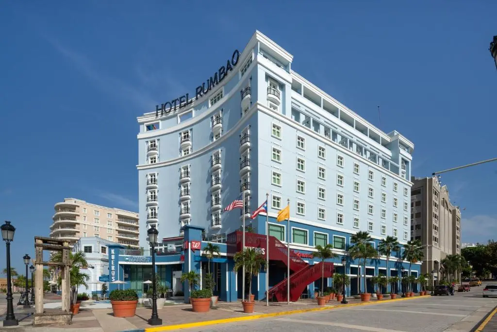 Hotel Rumbao's tall white building with blue trim and a red entrance stair, palm trees and flags out front under a blue sky