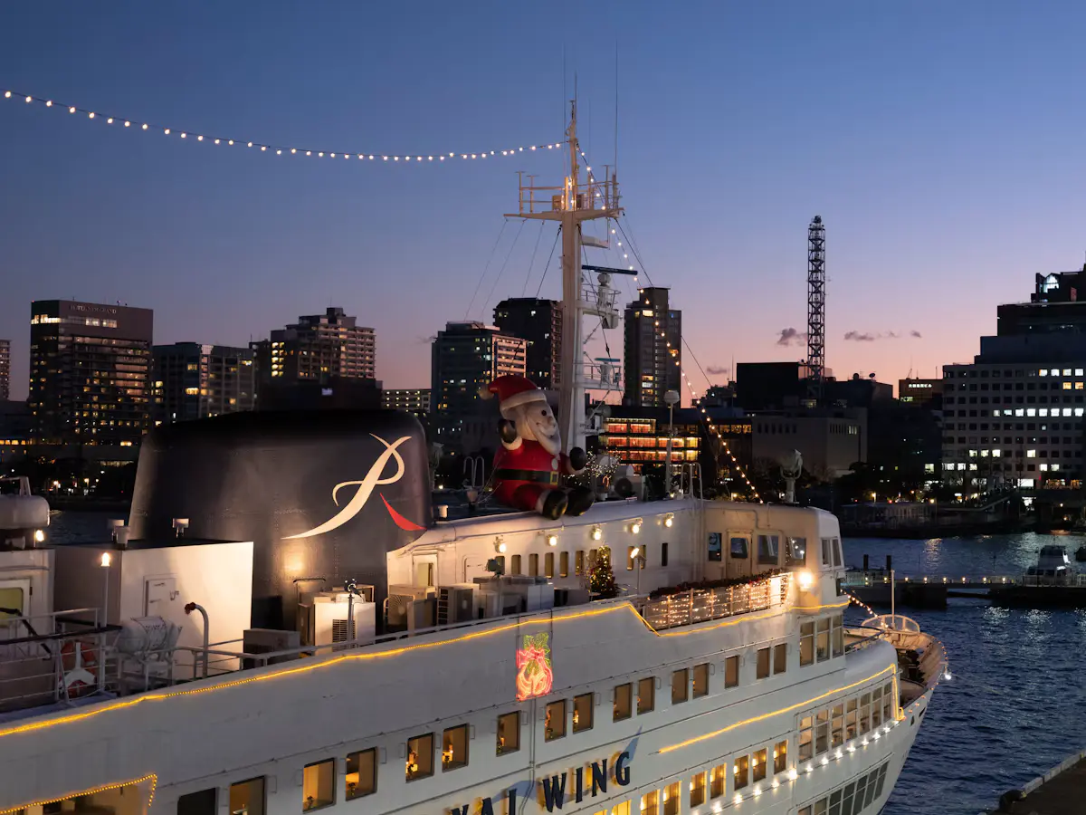 White ship strung with lights and a Santa figure on its upper deck at dusk, moored beside Yokohama's harbourside buildings under a blue and pink sky