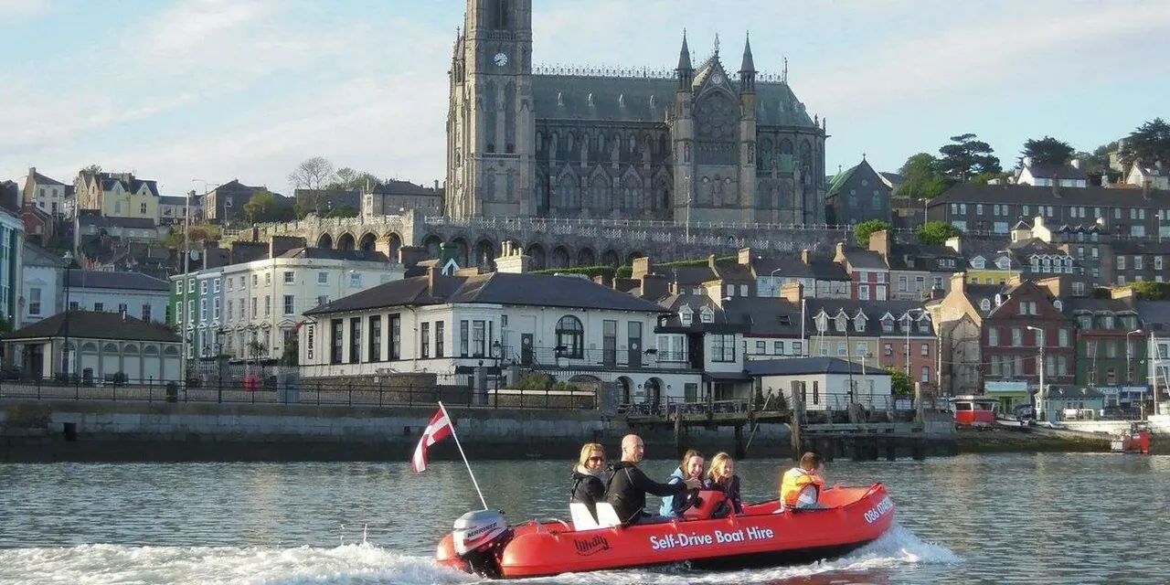 A small red boat carrying passengers across the harbour in front of Cobh's waterfront and cathedral