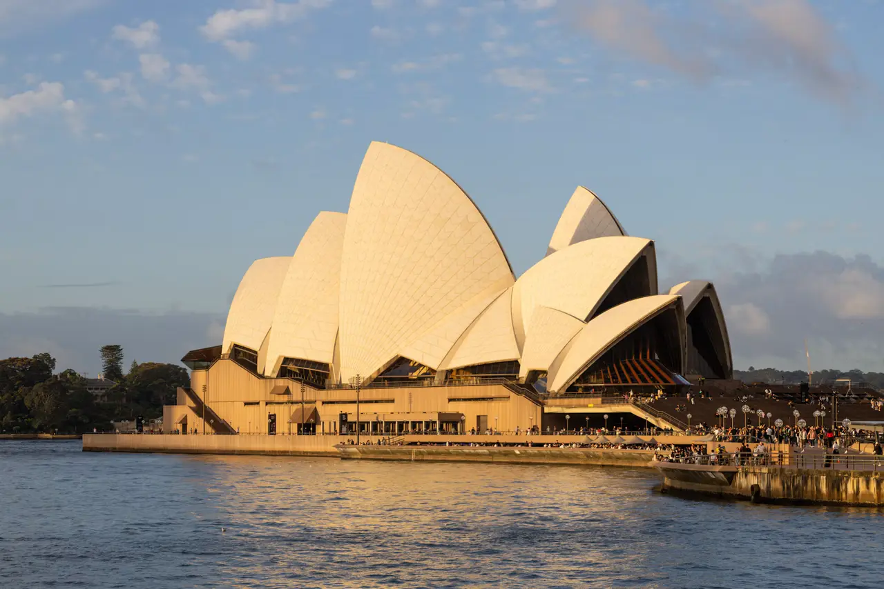 The white sails of the Sydney Opera House glowing gold in late sun beside calm blue harbour water, with soft clouds above