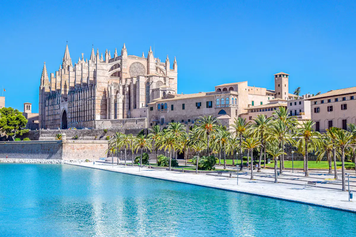 Palma Cathedral in honey-coloured stone behind palm trees and the turquoise water of the Parc de la Mar pond under a clear blue sky