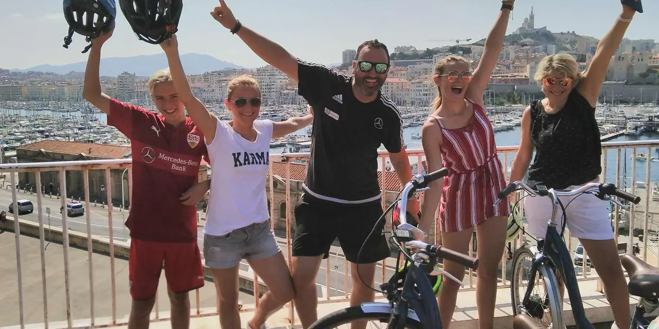 Three people holding bike helmets in the air on a terrace above Marseille's Old Port marina
