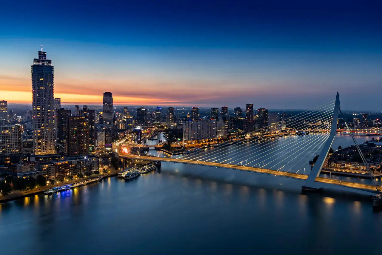 The Erasmus Bridge and the Rotterdam skyline from above at dusk, the bridge's cables lit and the sky fading from deep blue to orange over the river