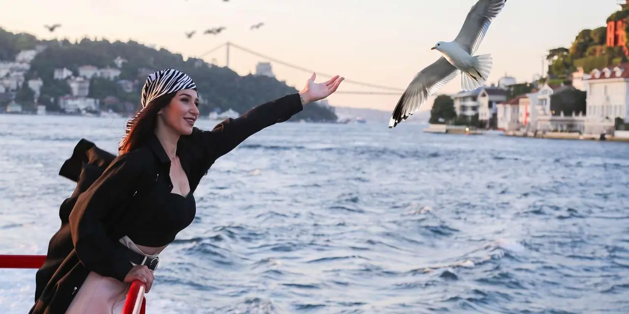 A woman in a headscarf waving at gulls from a boat's deck, with the green Bosphorus shore and a suspension bridge behind