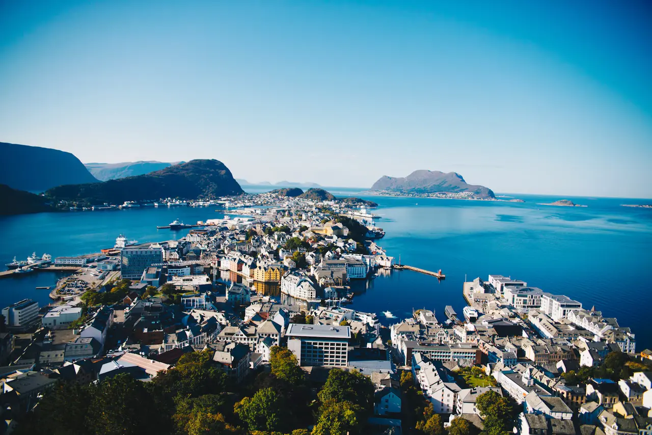 Ålesund from the Aksla viewpoint on a clear summer day, the town's rooftops spread across islands in a deep-blue sea with mountains and offshore islands behind