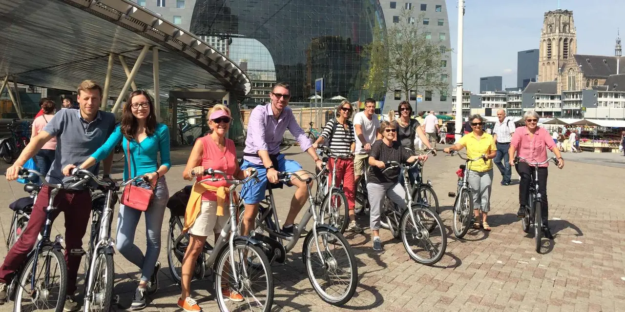 A group of about a dozen cyclists posing on their bikes on a paved square, with the Markthal's glass arch and a church tower behind them on a sunny day