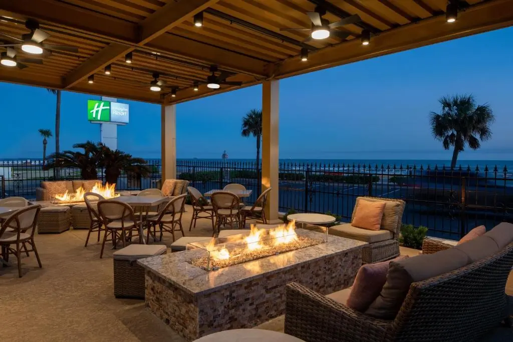 Fire-pit lounge on the Holiday Inn Resort Galveston's covered patio at dusk, with the seawall road and Gulf behind it