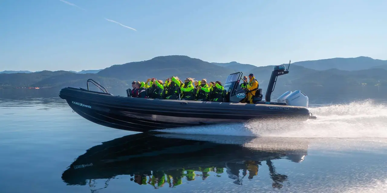 A dark inflatable boat planing across calm blue water, full of passengers in green and yellow flotation suits, with mountains behind