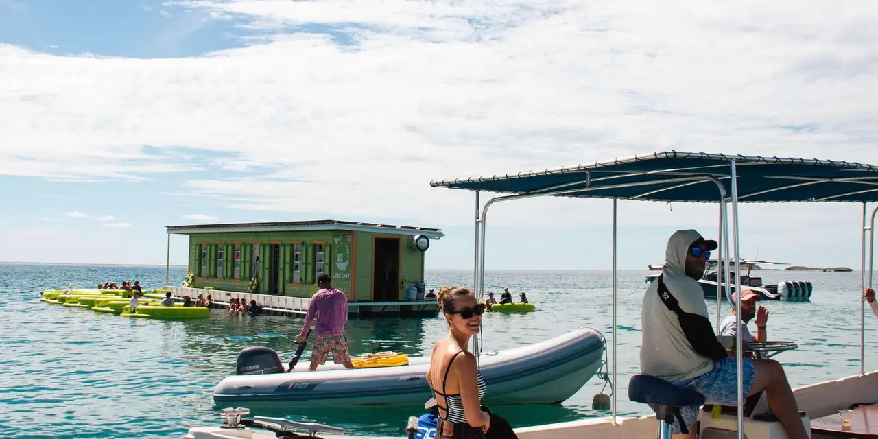 A small green floating bar anchored in a calm bay, with swimmers nearby and a canopied tour boat and dinghy in the foreground