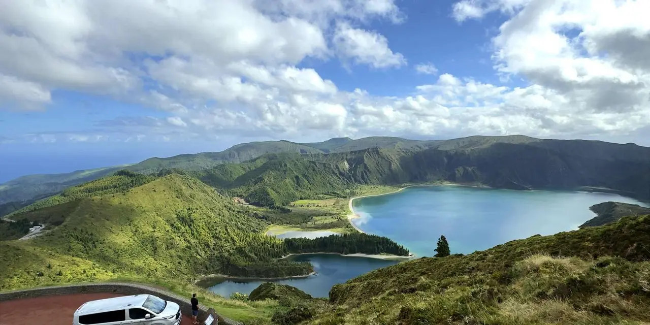 A white van parked at a roadside viewpoint above a blue crater lake ringed by green hills